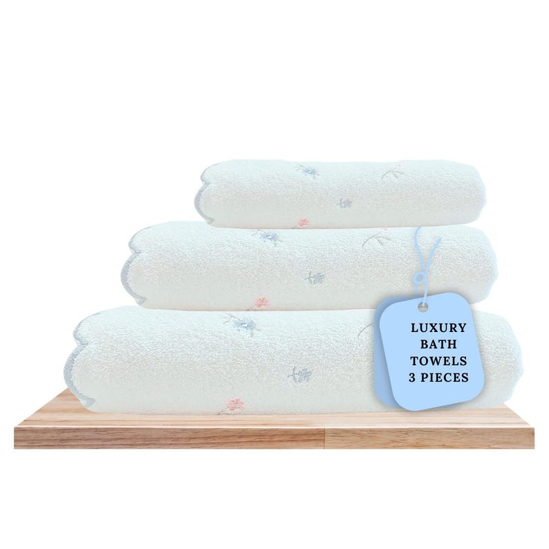 May include: Three white bath towels with a floral pattern stacked on a wooden shelf. A blue tag with the text "Luxury Bath Towels 3 Pieces" is attached to the towels.