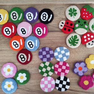 May include: A collection of circular tufted coasters featuring various designs including billiard balls with the number eight, checkered patterns, flowers, a strawberry, a ladybug, a frog, a mushroom, and green leaves in assorted colors.