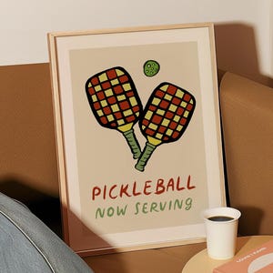 May include: A framed print with a beige background featuring two red and yellow pickleball paddles with a green ball above them. The text "Pickleball Now Serving" is written in black below the paddles.