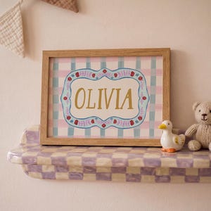 May include: A framed print with the name "OLIVIA" in gold lettering, set against a pink and blue plaid background. The frame is light brown. A ceramic duck and a teddy bear sit on a checkered shelf below the print. A decorative banner hangs above.