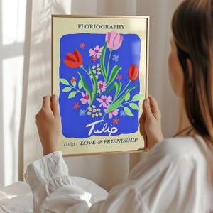 A person is holding up a framed poster with a colorful illustration of flowers, including tulips, against a blue background.