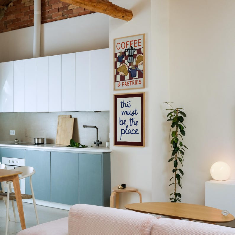 May include: A contemporary kitchen featuring white and blue cabinets, a stainless steel sink, and two framed art prints. One print reads "Coffee & Pastries" and the other says "this must be the place."