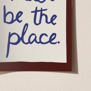 May include: A framed print featuring the phrase "be the place" in blue script on an off-white background. The print is framed with a dark red border, offering a bold contrast. The design is simple and contemporary.