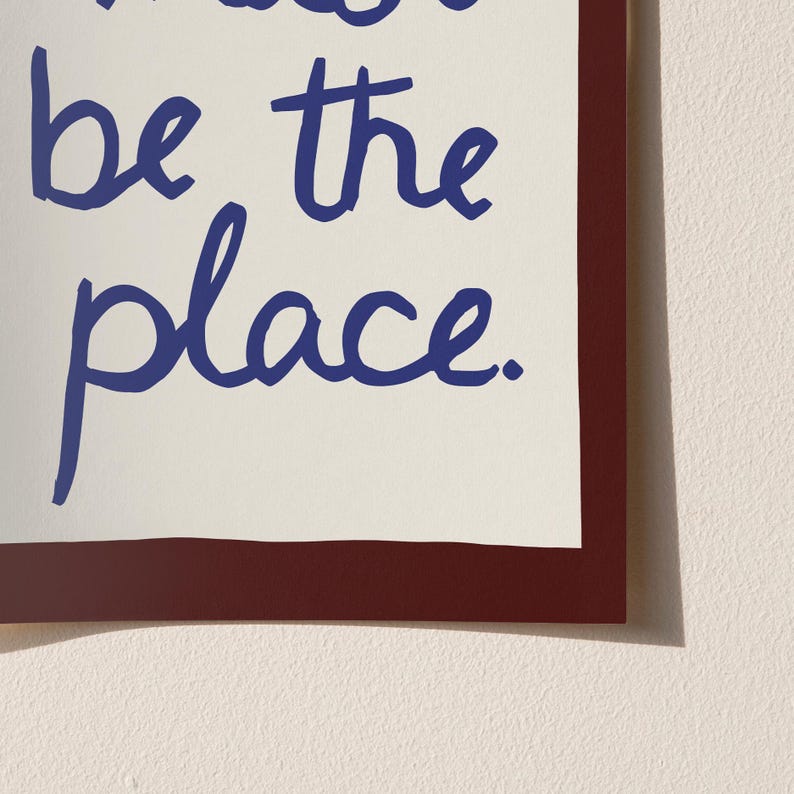May include: A framed print featuring the phrase "be the place" in blue script on an off-white background. The print is framed with a dark red border, offering a bold contrast. The design is simple and contemporary.
