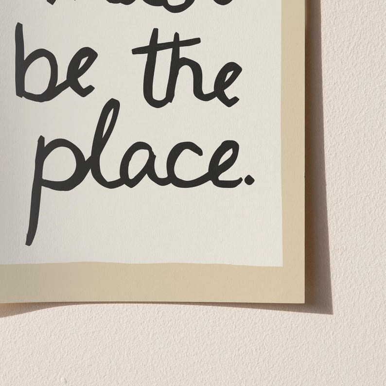 May include: A framed print featuring the phrase "be the place" in black cursive lettering. The text is set against a cream backdrop, framed by a light beige border. The print is mounted on a light beige wall.