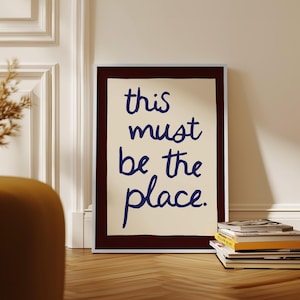 May include: A framed art print with the words "this must be the place" in navy blue script on an off-white background. The frame is dark brown with a white border. A stack of books sits on the wooden floor.