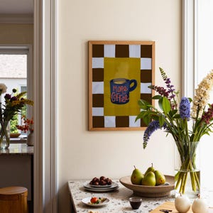 a cozy kitchen scene with a table set for breakfast. On the table, there is a bowl of fruit, including pears, and a vase of flowers. Above the table, there is a framed picture with a coffee mug and the text "MORE COFFEE" written on it. The kitchen also has a window and a chair visible in the background.