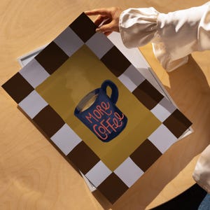 A person's hand is holding a framed print with a graphic of a coffee mug and the text "More Coffee" written on it.