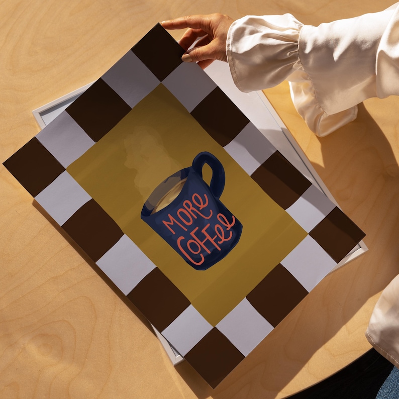 A person's hand is holding a framed print with a graphic of a coffee mug and the text "More Coffee" written on it.