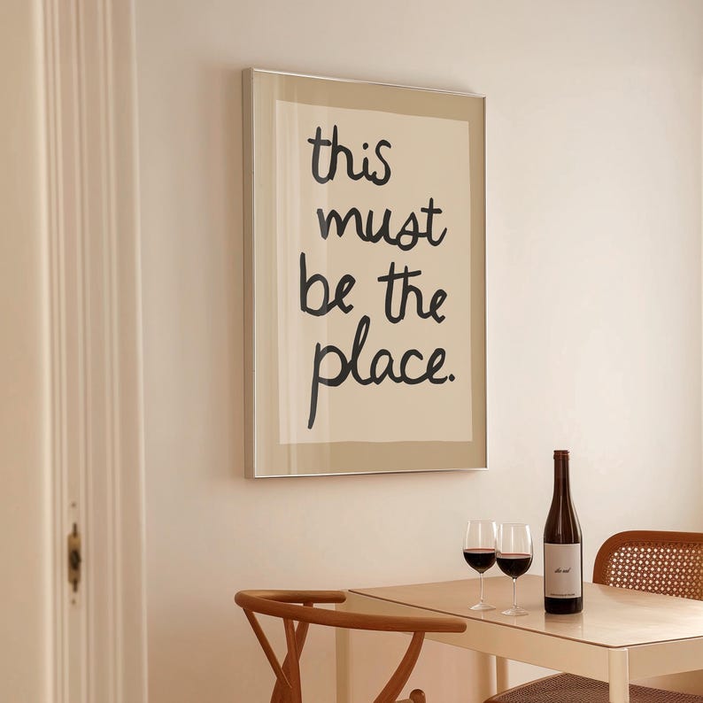 May include: A framed print with the words "this must be the place" in black script hangs above a table set for a meal. A bottle of wine and two glasses of red wine sit on the table, with a wooden chair nearby. The room has a neutral colour palette.