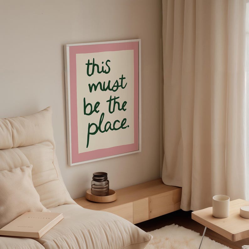 May include: A framed art print with the text "this must be the place" in green script on a cream background, framed in pink and white. The print hangs on a white wall. A glass jar and a small table are also visible.