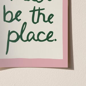 May include: A framed print featuring the words "be the place" in dark green script. The text is set against an off-white background, with a pink border. The print is displayed against a light beige wall.
