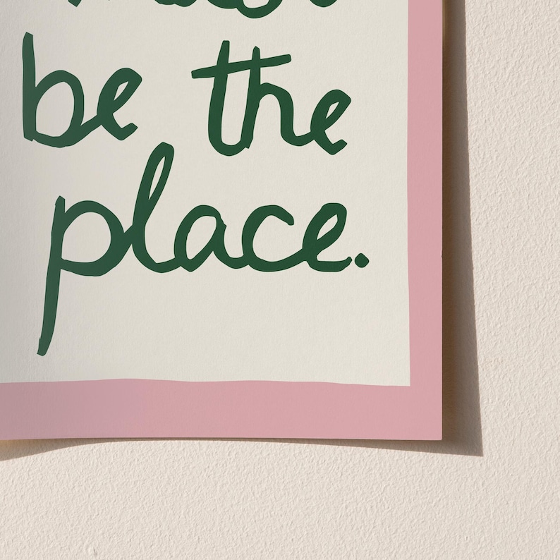 May include: A framed print featuring the words "be the place" in dark green script. The text is set against an off-white background, with a pink border. The print is displayed against a light beige wall.