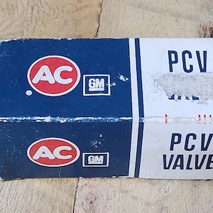 May include: A vintage AC Delco PCV valve box. The box is divided into blue and white sections with the AC logo in a red circle. The text "PCV VALVE" is printed on the white section. The box is on a wooden surface.