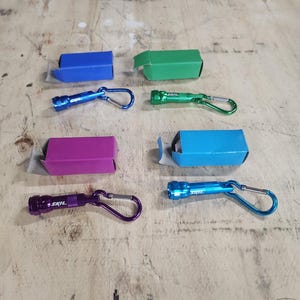 May include: Six small keychain flashlights in different colors, including blue, green, purple, and turquoise. Each flashlight has a carabiner clip and the word "SKIL" printed on the side.