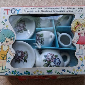 May include: A miniature porcelain tea set in a rectangular box featuring illustrations of two children. The set includes a teapot, cups, and saucers decorated with floral patterns. The box text reads: CAUTION: Not recommended for children under 8 years old. Contains breakable china.