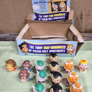 May include: A collection of vintage "Egg-Heads" figurines, each with a unique painted face and character. The display box reads "The Funny Egg-Dorables of Pigeon-Hole Apartments" and features cartoon graphics. The figurines are in various colors.