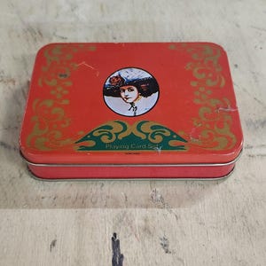 May include: A vintage red playing card set tin box. The box features a portrait of a woman in a hat and the words "Playing Card Set". Gold filigree designs adorn the edges. The box is rectangular and made of metal.