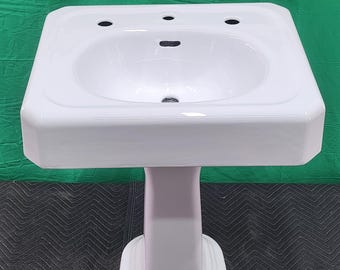 May include: A white porcelain pedestal sink with a rectangular basin and an oval bowl. The sink has three faucet holes and a central drain. The pedestal base is a matching white color and has a rounded, stepped design. The sink is clean and appears new.
