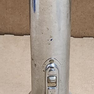 May include: A vintage silver flashlight with a cylindrical body and a circular base and top. The switch is rectangular and reads "MADE IN U.S.A.". The top of the flashlight has the word "Trail-Lite" embossed.