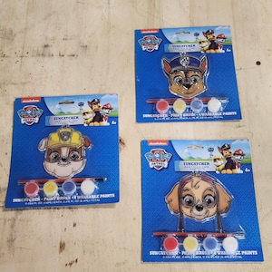 May include: Three Paw Patrol character suncatchers with paint and a paintbrush. Each suncatcher features a different character: Chase, Rubble, and Skye. The paint pots are red, yellow, blue, and white. The packaging says "SUNCATCHER PAINT YOUR OWN CHARACTER" and "4+."