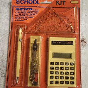 May include: A school kit with a yellow calculator, compass, ruler, protractor, and pencil. The packaging is orange and white with the text "SCHOOL KIT" and "aurora AK-100".