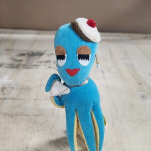 May include: A blue plush octopus wearing a white hat with a red cherry on top. The octopus has yellow accents on its tentacles and is holding a white paper object in its hand.