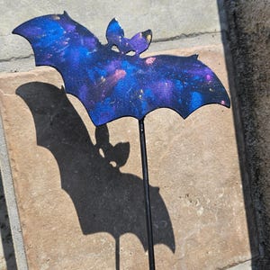 May include: A purple and blue bat silhouette with a galaxy-like design. The bat is attached to a black metal stake.