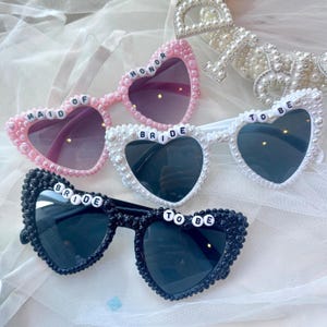 Puede incluir: Gafas de sol en forma de corazón en rosa, blanco y negro, adornadas con cuentas similares a perlas. El par rosa dice "MAID OF HONOR", el par blanco dice "BRIDE TO BE", y el par negro dice "BRIDE TO BE". Las lentes están teñidas.