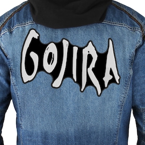 Könnte beinhalten: Ein gestickter Patch in Schwarz und Weiß mit dem Wort "GOJIRA" in einer stilisierten Schriftart. Der Patch ist auf eine blaue Jeansjacke genäht.
