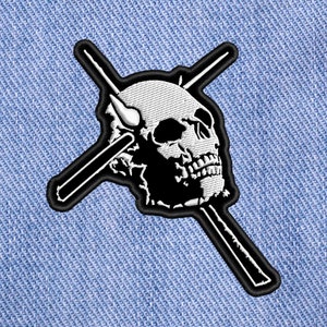 May include: White embroidered skull with two crossed black drumsticks on a blue denim background.