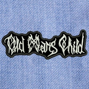 Può includere: Una toppa ricamata in bianco e nero con la scritta "Old Man's Child" in un font gotico. La toppa rettangolare ha un bordo nero ed è posta su uno sfondo di denim blu.