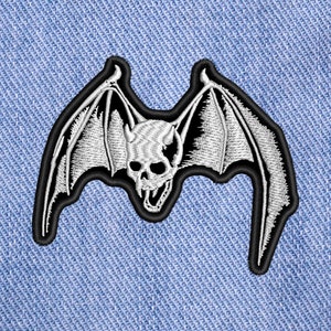 Könnte beinhalten: Schwarz-weiß gestickter Aufnäher mit einem Schädel mit Fledermausflügeln. Der Schädel hat Hörner und ein zahniges Grinsen.