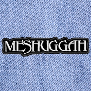 Op de afbeelding: Een rechthoekige geborduurde patch met het woord MESHUGGAH in witte gestileerde letters met een zwarte rand, gecentreerd op een lichtblauwe spijkerstof achtergrond met een zichtbare diagonale keperbinding in het gehele materiaal.