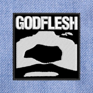 May include: Embroidered patch with the word "GODFLESH" in white, block letters above a white and black abstract design. The patch is square with a black border, set against a light blue denim background.