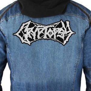 Könnte beinhalten: Eine blaue Jeansjacke mit schwarzer Kapuze und einem großen, schwarz-weißen Aufnäher auf dem Rücken. Der Aufnäher zeigt das Wort "CRYPTOSY" in einer weißen, gotischen Schrift. Die Jacke hat braune Akzente.