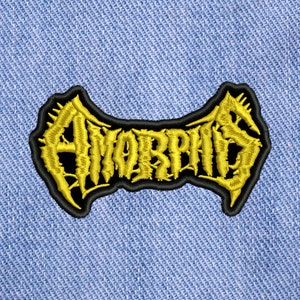 Puede incluir: Un parche bordado amarillo con la palabra "AMORPHIS" en contorno negro. El parche está sobre un fondo de mezclilla azul.