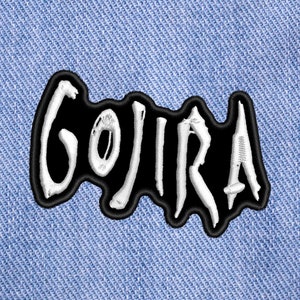 Könnte beinhalten: Weiß gestickter "GOJIRA"-Text auf einem schwarzen Patch. Der Patch befindet sich auf einem blauen Jeansstoff.