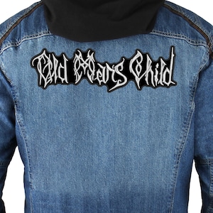 Op de afbeelding: Een spijkerjasje met een zwarte capuchon en een grote patch op de rug. De patch toont de tekst "Old Man's Child" in een gotisch lettertype met witte letters en een zwarte rand. De jas heeft bruine accenten.