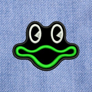 以下が含まれることがあります： 黒と緑の刺繍でできたカエルのワッペン。目は白く刺繍されている。
