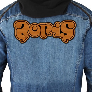 Puede incluir: Una chaqueta vaquera marrón con un parche bordado marrón que dice "Boris" en la espalda.