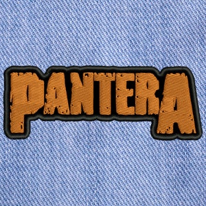 May include: An embroidered patch featuring the word PANTERA in a distressed, textured orange font with a thick black outline. The patch is stitched onto a light blue denim fabric background with a visible diagonal twill weave pattern.