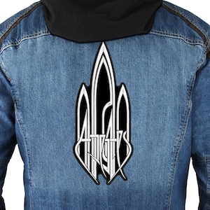 Puede incluir: Una chaqueta vaquera con un parche negro y blanco en la espalda. El parche presenta un diseño estilizado con la palabra "Atticus" en blanco.