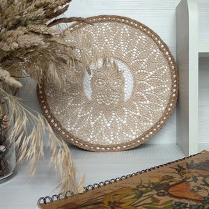 May include: A round, beige crocheted wall hanging with a white center design. The hanging is on a white shelf with a book and dried grasses in a vase.