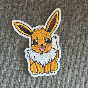 May include: Embroidered patch featuring a cartoon Eevee character. The patch has a yellow body, white fur, and large eyes. The ears are yellow with brown accents. The character is smiling with a pink tongue. The patch is on a gray background.
