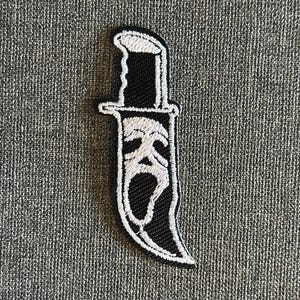 May include: Black and white embroidered patch featuring a knife with a Scream mask design. The patch has a black handle and blade with a white outline and a white Scream mask design on the blade.