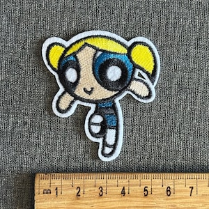 May include: Embroidered patch featuring the character Bubbles from The Powerpuff Girls. The patch has a white border, with Bubbles' face and body in yellow, blue, black, and white. A wooden ruler with millimeter markings is placed below the patch.