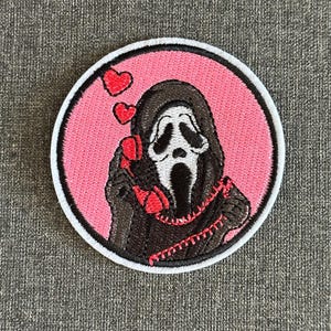 Könnte beinhalten: Gestickter Aufnäher mit der Ghostface-Maske aus der Scream-Filmreihe. Das Design umfasst einen rosa Hintergrund, drei rote Herzen und ein rotes Telefonkabel. Der Aufnäher ist kreisförmig mit weißem Rand und schwarzer Umrandung.