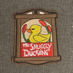 Peut inclure: Écusson brodé avec un canard jaune dans un cadre marron, avec le texte "The Snuggly Duckling" en rouge. Le canard est entouré d'éléments rouges et verts, sur fond gris.