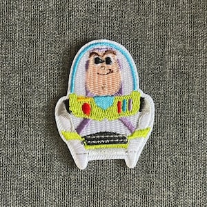 Può includere: Toppa ricamata con Buzz Lightyear di Toy Story. La toppa è bianca con un casco blu, dettagli gialli e verdi e un design in bianco e nero nella parte inferiore. Il volto del personaggio è visibile all'interno del casco.